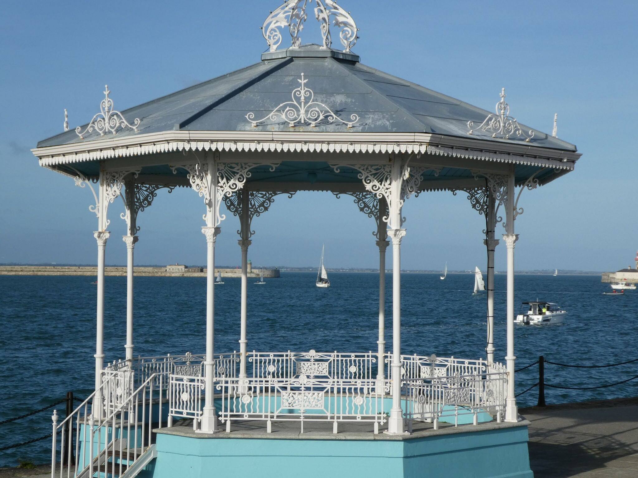 A pleasure yacht can be seen framed by the old white iron work of the band stand at Dun Laoghaire against a bright blue sky and deep blue water.