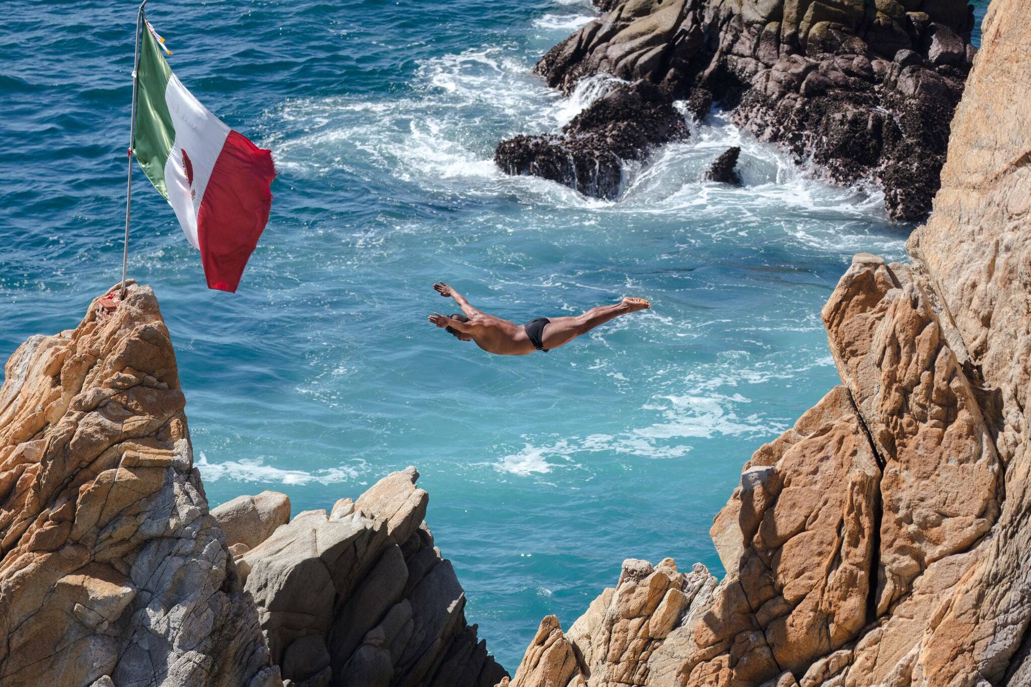 "La Quebrada, Acapulco/Mexico - December 2nd, 2018: A La Quebrada Cliff Diver courageously jumps into the sea below near a Mexican flag.