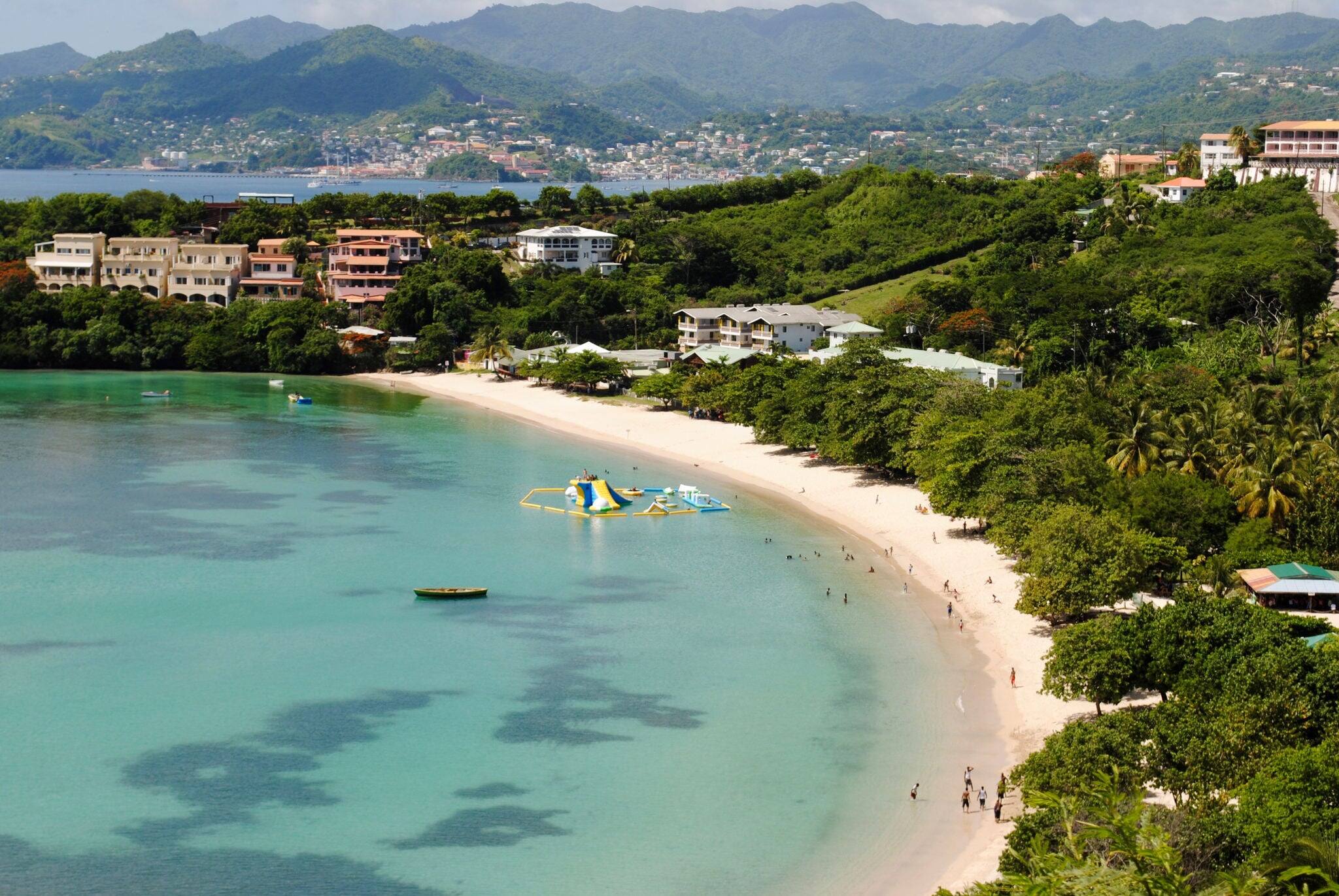 Grenada, south coast, white sand beach with floating play area in shallow water, small boats and people and the city of St. George's in the background.