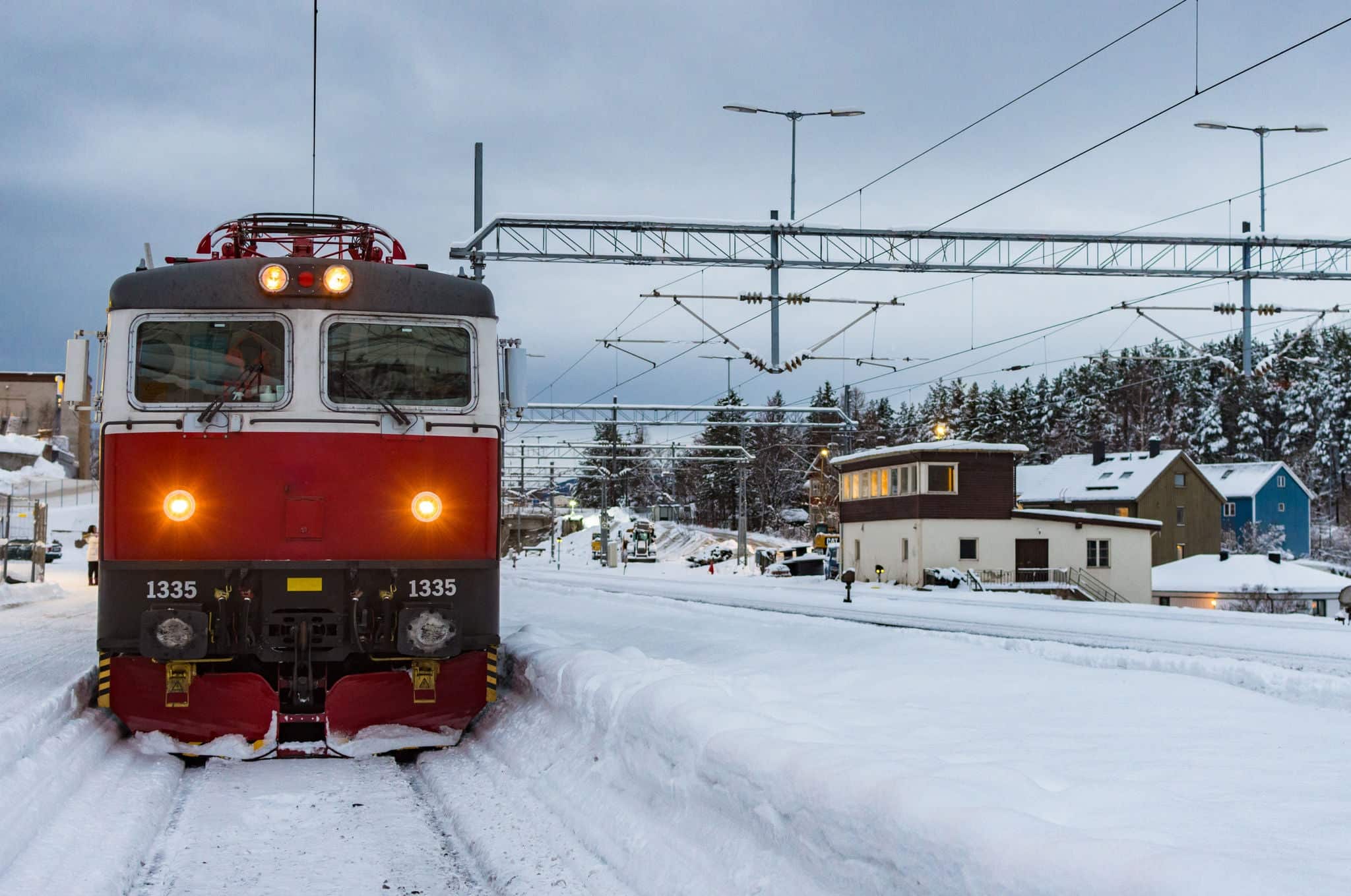 Tren rojo en una estación de tren nevada y fría junto a la plataforma, la estación y los árboles en el fondo.