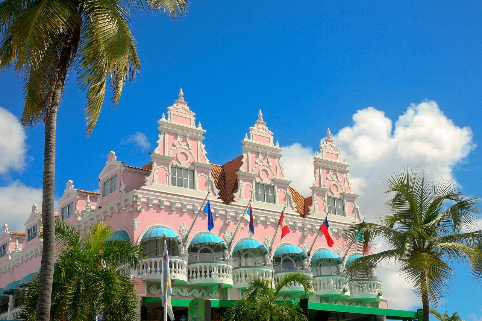 The pink Dutch style Royal Plaza with two palm trees at the side. It is set off with white clouds and blue sky. This building is an icon for Oranjestad.