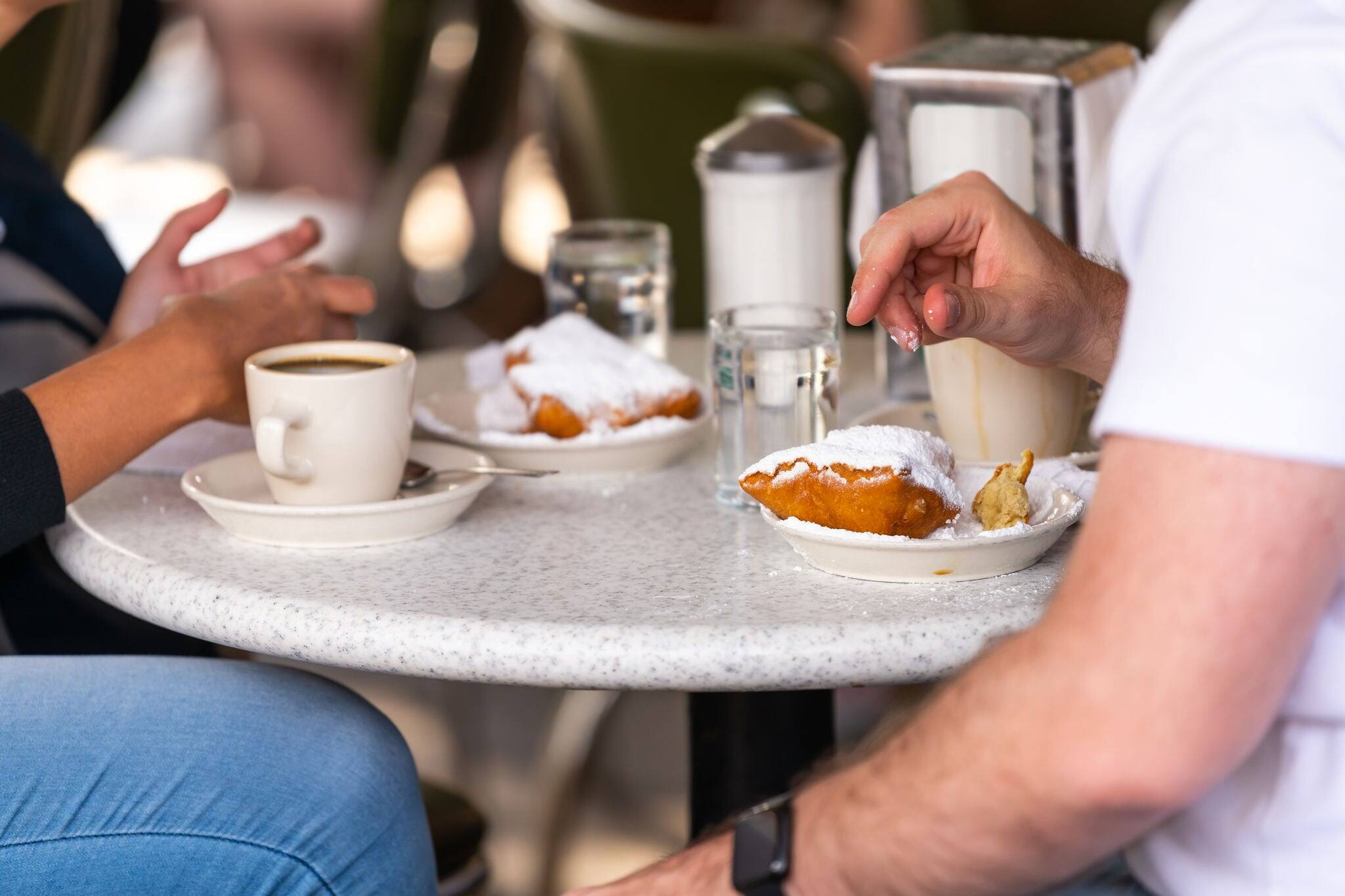 Closeup of couple sitting outside at sidewalk cafe by table drinking chicory coffee and eating deep fried beignet donut powdered with sugar