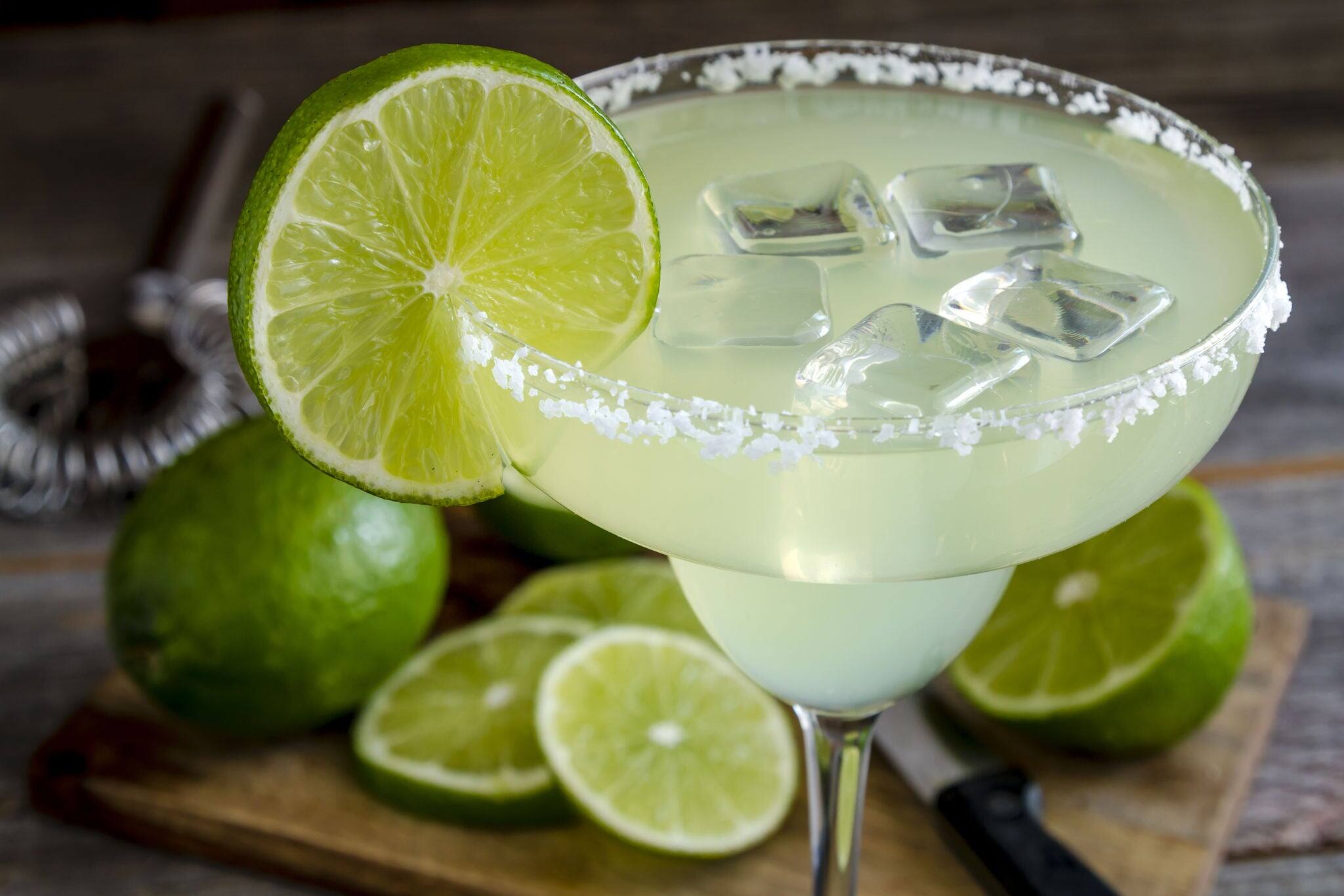 Close up of classic lime margarita cocktail with sliced and whole limes sitting on wooden cutting board